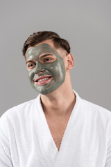 smiling young man with clay mask in white bathrobe looking away isolated on grey