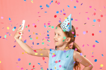 Cheerful little girl celebrates birthday. The child holds the phone, takes a selfie in the rain of confetti. Closeup portrait on pink background.