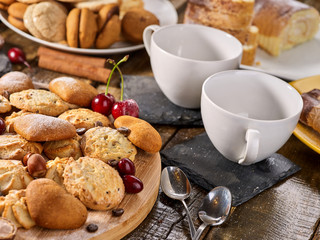 Oatmeal cookies and sand chocolate cake with cherry berry and crispy wafer rolls with cream on cutting board on wooden table in rustic style. Still life served with cups and dessert spoons. Useful for