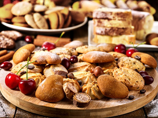 Oatmeal cookies and sand chocolate cake with cherry berry and crispy wafer rolls with cream on cutting board on wooden table in rustic style. Problems with excess weight. Sunlight in kitchen.