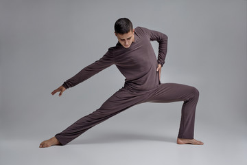 Photo of an athletic man ballet dancer dressed in a gray tracksuit, making a dance element against a gray background in studio.