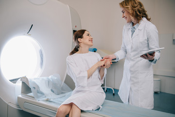 smiling radiologist holding hands with happy woman sitting on computed tomography scanner bed