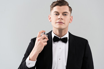 front view of confident man in formal wear applying perfume isolated on grey