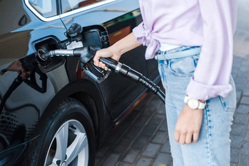 Obraz premium cropped view of woman holding fuel pump while refueling black car with benzine