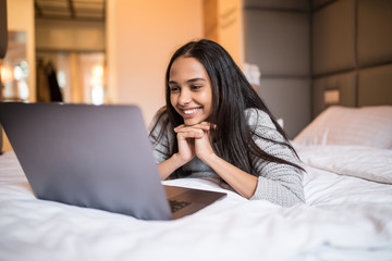 Young happy woman with laptop lying on bed at home. Lazy morning