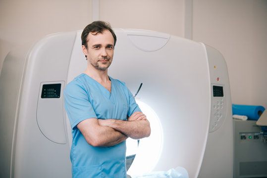 smiling  radiologist standing near computed tomography scanner with crossed arms and looking at camera