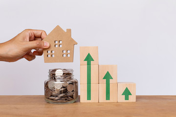 Hand putting house model on money coins in glass jar withWooden cube stack a staircase and arrows...