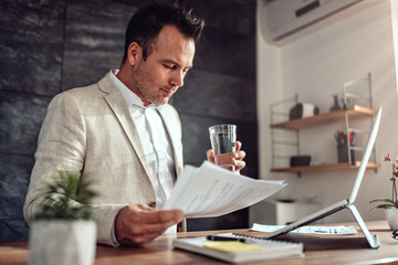 Businessman doing paperwork in his office and holding glass of water