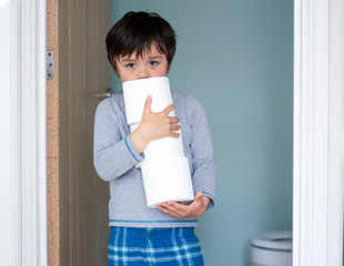Portrait of cute little boy holding toilet roll standing in front of toilet, Child wearing pajamas carrying a stack of toilet paper with bored face, Kid helping parent doing house work
