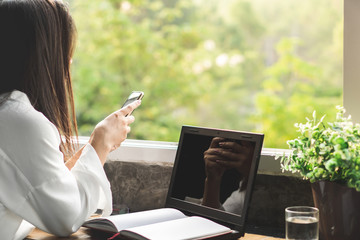 Asian business woman working from home office hand holding smart phone and laptop on desk with nature view next to the window