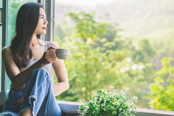 beautiful Asian woman sitting next to the window drinking coffee and relaxing with beautiful nature view  - Powered by Adobe