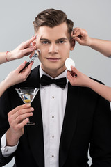 cropped view of makeup artists doing makeup for man in formal wear holding glass of cocktail isolated on grey