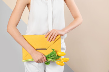 Stylish woman with clutch and spring flowers against color background, closeup