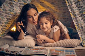 Mother and daughter are sitting in a teepee tent, reading stories with the flashlight. Happy family. © nazarovsergey