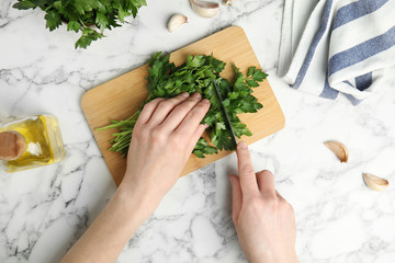 Woman cutting fresh green parsley on wooden board at marble table, top view