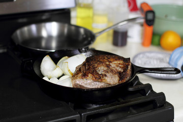 Sirloin steak frying in a cast iron pan with onions on the stove top.