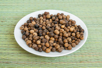 A lot of allspice berries on a white saucer on a green table mat made of natural plant fibers. Natural food spices and seasonings.