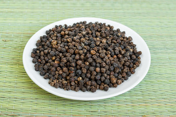 A lot of black pepper grains on a white saucer on a green table mat made of natural plant fibers. Natural food spices and seasonings.