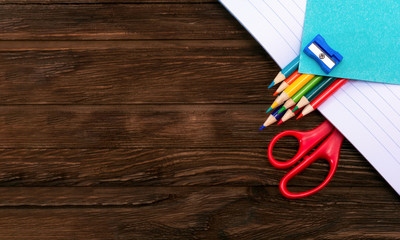 Obraz premium Colored pencils, sharpener, scissors and sheets for notes in an open notebook on a dark brown wooden surface. Flat lay. Back to school. Education. Office tool. Empty place for text.