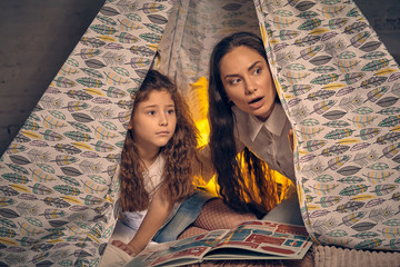 Mother and daughter are sitting in a teepee tent with some pillows and reading stories. Happy...