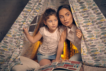 Mother and daughter are sitting in a teepee tent with some pillows and reading stories. Happy family.