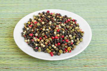 Peppercorn melange. White, black, red and green pepper grains on a white saucer on a green table mat made of natural plant fibers. Natural food spices and seasonings.