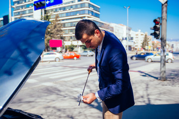 A handsome businessman wearing blue blazer lifting up the hood of his car and checking the oil level on a sunny day parked on a busy city boulevard.