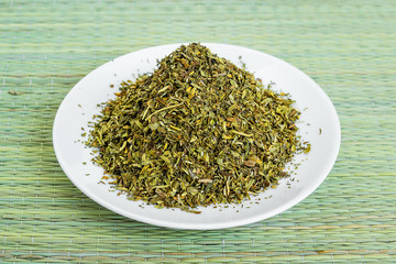 Fototapeta na wymiar A heap of dry tarragon or estragon flakes on a white saucer on a green table mat made of natural plant fibers. Natural food spices and seasonings.