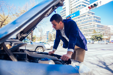 A handsome businessman wearing blue blazer lifting up the hood of his car and checking the oil level on a sunny day parked on a busy city boulevard.