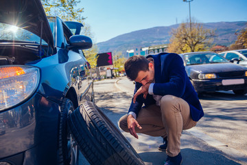 Angry businessman trying to change his flat tire with a socket wrench while crouching down next to his car.