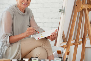 Smiling mature woman sitting and relaxing during painting a picture on canvas in art studio