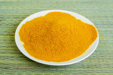 A lot of turmeric or curcuma powder on a white saucer on a green table mat made of natural plant fibers. Natural food spices and seasonings.