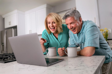 Smiling and laughing retired seniors video chatting with family on internet with laptop in home...