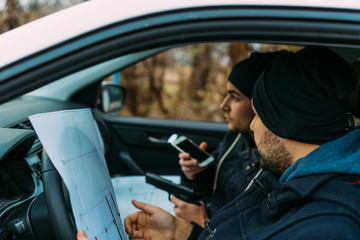 Two armed bandits sitting in a car planning their next robbery, while counting on a stopwatch the time they have for the robbery, showing off their guns and pointing at the blueprint.