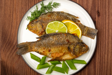 Grilled Crucian fish decorated with lemon slices with lime and fresh herbs. Ready meal on a plate, wooden background