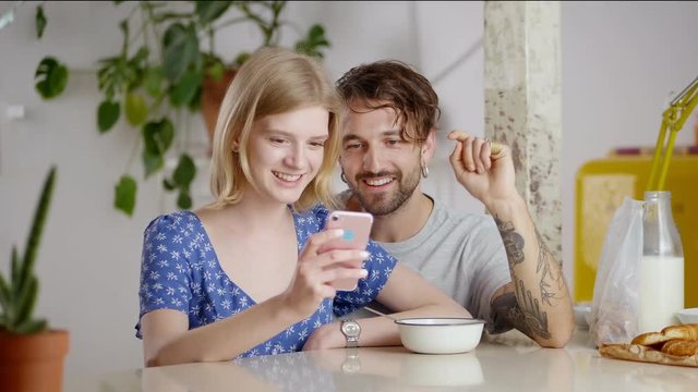 Portrait of blonde girl holding smartphone guy scrolling news feed in social application, cute mixed raced couple enjoying to spend free time together during cozy brunch leaning at bar table in loft
