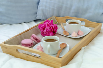 Breakfast in bed for two. Wooden tray with coffee, macaroons and Bizet. Decoration pink peony. Beautiful natural light from the window.