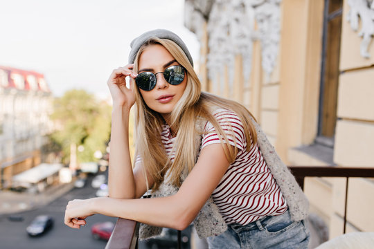 Lovely girl with pale skin posing in dark glasses while chilling at balcony. Close-up photo of adorable young lady in sport hat and summer t-shirt relaxing on terrace and listening city sounds.