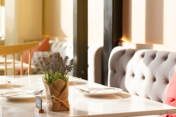Dining table in restaurant with dishes set and napkin flower pot and table number with gray long sofa and wooden chair in evening time.