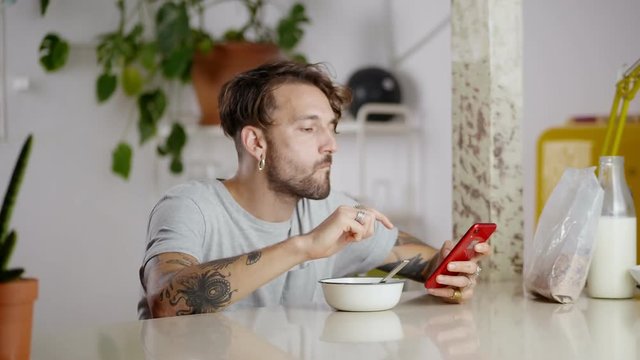 Side portrait of cute man eating breakfast and using red gadget online in social network, bottle of milk and pack of corn flakes on kitchen table, smiling guy sitting with smartphone white wall around