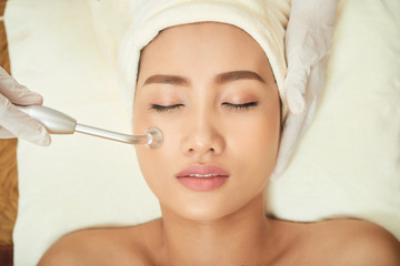 Asian beautiful woman relaxing with eyes closed while cosmetologist in protective gloves doing ultrasound procedure for her face