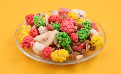 Mix nuts and dried fruits. Cleaned kernels of peanut, walnut, assortment of peanut in colored sugar, candied fruits, raisin and banana chips in glass plate on yellow background. Selective focus.