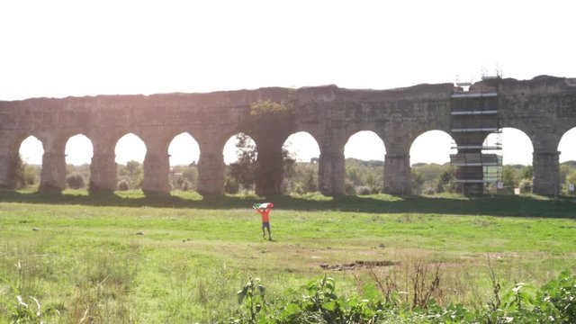 Young man in orange sportswear wearing italian flag as cape exults running in front of ancient roman aqueduct in parco degli acquedotti in rome at sunset