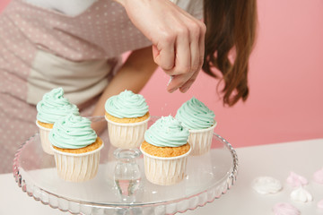 Close up cropped housewife female chef cook confectioner or baker in apron white t-shirt decorating cake cupcake at table isolated on pink pastel background in studio. Mock up copy space food concept.