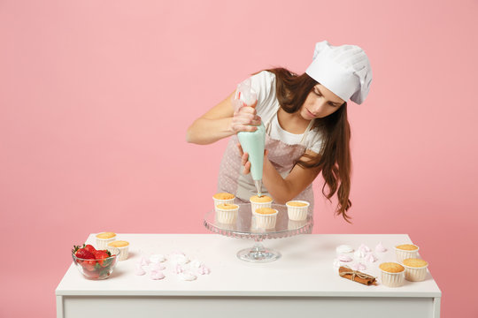 Housewife female chef cook confectioner or baker in apron white t-shirt, toque chefs hat decorating cake cupcake at table isolated on pink pastel background in studio. Mock up copy space food concept.