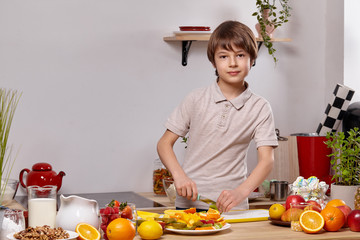 Cute little boy with brown hair is cooking at a kitchen against a white wall with shelves on it.