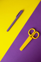 Yellow kid's stationery scissors on purple background, purple pen on yellow background. Yellow and purple background halve image diagonally. Back to school concept. Top view of office supplies.