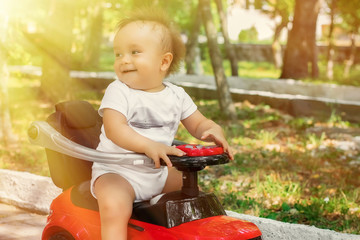 Little cheerful infant in white shirt sitting half turned back on red push car in park or garden in sun light. Happy active childhood and family vacation concept. Toned image.
