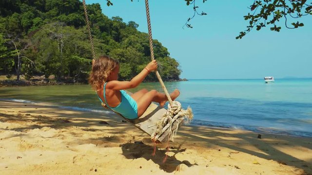 Cute blonde curly kid girl in blue swimsuit is swinging on a sandy beach by the ocean in tropical island at sunny warm day.