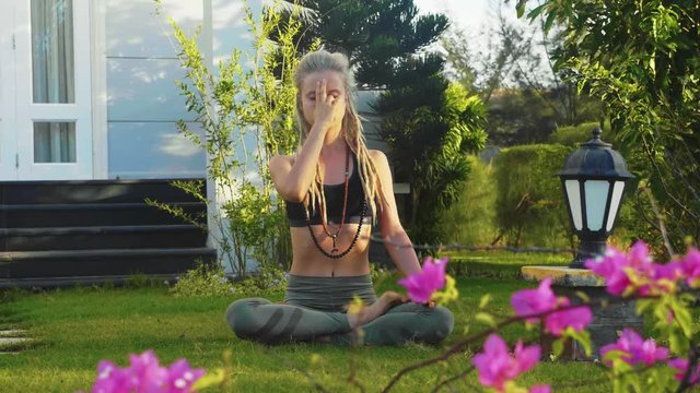 Pranayama yoga breathing practice of blonde young woman with dreadlocks sitting in lotus pose with hand fingers on her forehead on courtyard of her house.